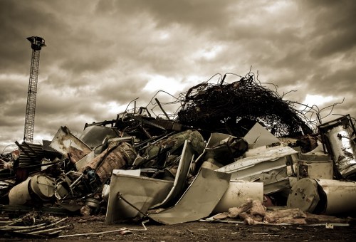 A large pile of discarded metallic objects, including white and grey appliances, scrap metal, and tangled wires, is situated outdoors on a dirt surface. The appliances appear old and broken, with some showing rust and corrosion on their surfaces. The pile is uneven, with items stacked haphazardly, and is topped by a mass of tangled dark debris or wire mesh. In the background, a tall industrial or communication tower is visible to the left, under a cloudy sky with dark overcast clouds suggesting overcast weather. The scene appears to be in an open, possibly industrial or waste disposal area, with no vegetation visible, and the overall atmosphere is subdued and utilitarian, reflecting typical surroundings for rubbish removal in a London borough such as Elephant and Castle, with a focus on heavy metallic waste.