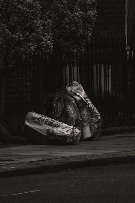 Two large black plastic bags filled with waste are placed on the pavement in front of a dark metal fence, with dense foliage partially visible behind the fence. The bags appear to contain household ru