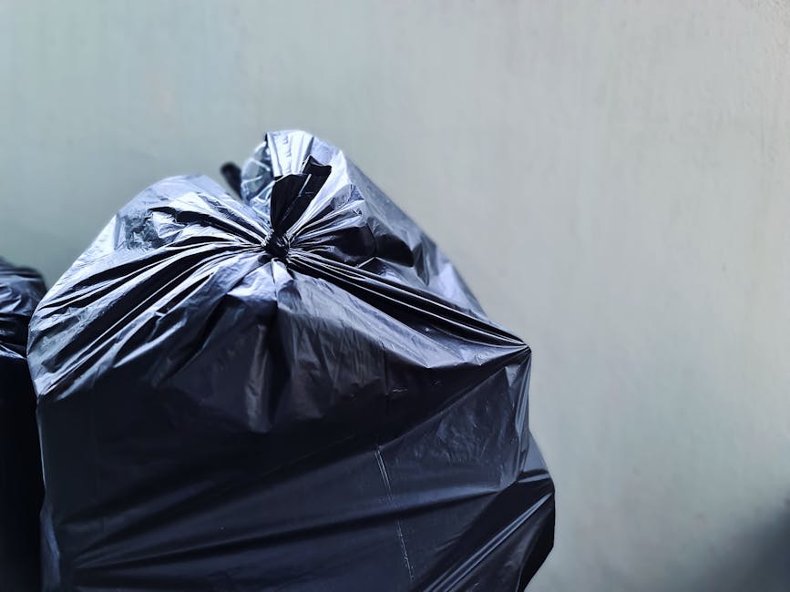 Two large black plastic bags filled with waste are placed on the pavement in front of a dark metal fence, with dense foliage partially visible behind the fence. The bags appear to contain household ru