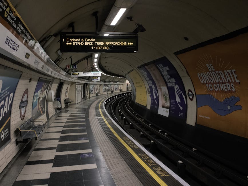 An underground train station platform with a curved track and a tiled floor featuring black, white, and grey tiles arranged in a checkered pattern. The platform is illuminated by a row of ceiling ligh