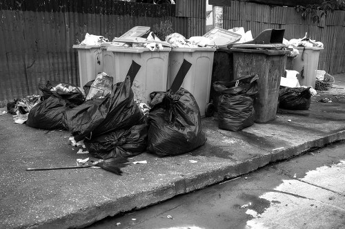 A collection of black plastic rubbish bags and various discarded items, such as paper and packaging, are piled on a concrete pavement in front of several large wheelie bins, which are situated against a wooden fence in an outdoor area. The wheelie bins include a light grey and a dark grey bin, with some lids open and filled with rubbish, while others are closed. The surrounding environment suggests a typical residential or commercial waste collection point in Elephant and Castle, with the bags and waste extending slightly onto the uneven pavement surface. Natural daylight illuminates the scene, highlighting the different textures of the plastic bags, paper, and the metal and plastic finishes of the bins. The image conveys a scene typical of rubbish removal services provided by Business Waste Removal Elephant and Castle, illustrating collection and disposal tasks in an urban setting, with attention to waste containment and environmental cleanliness in the local postcode area.